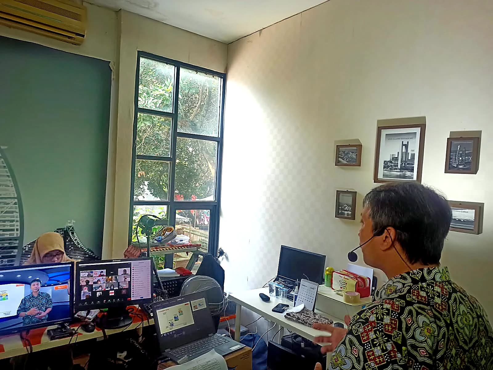 Trainer dengan headset memandu sesi online dari ruang kerja kantor, layar Zoom peserta terlihat