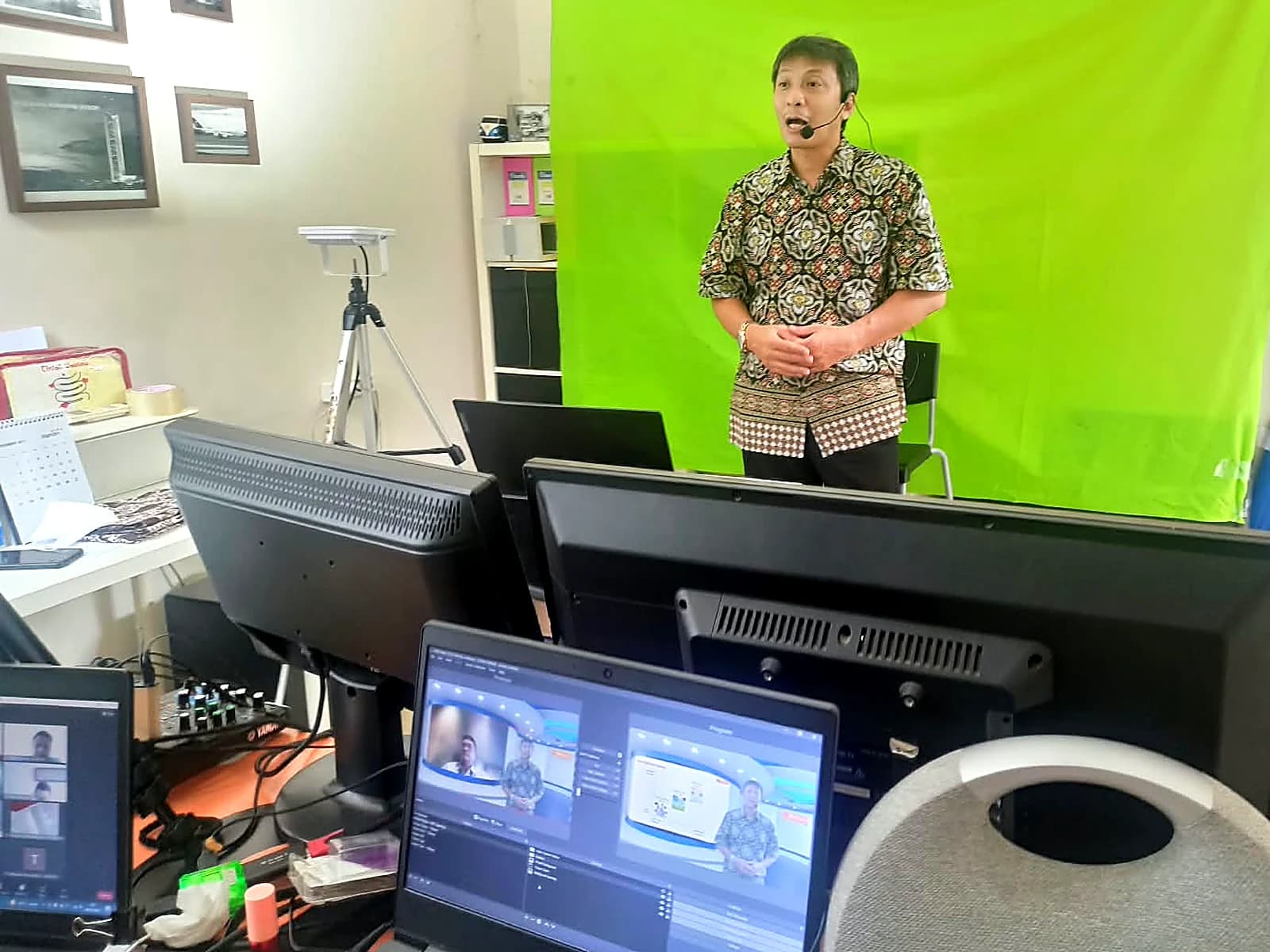 Trainer berdiri di depan green screen saat perekaman, dengan monitor produksi di latar depan